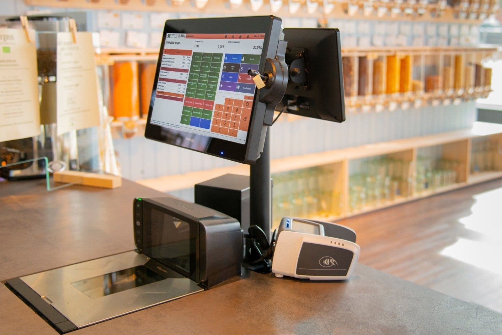 beePOS-Kasse mit Kundendisplay, Scanner und Kartenterminal vor den Abfüllstationen eines Unverpackt-Ladens